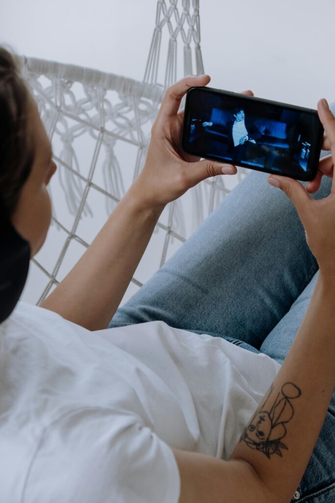 A woman in blue jeans and headphones watches a video on her smartphone while relaxing indoors.